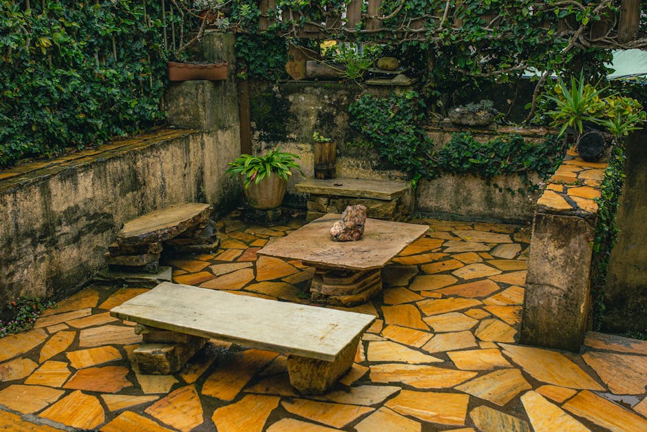 A small, private garden patio enclosed by weathered concrete walls, featuring a decorative stone floor with irregularly shaped amber and brown tiles. The space includes a low, rectangular stone table